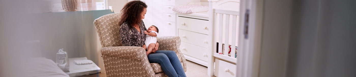 A mother sitting on a cosy nursering chair cradling her baby in their nursery room.